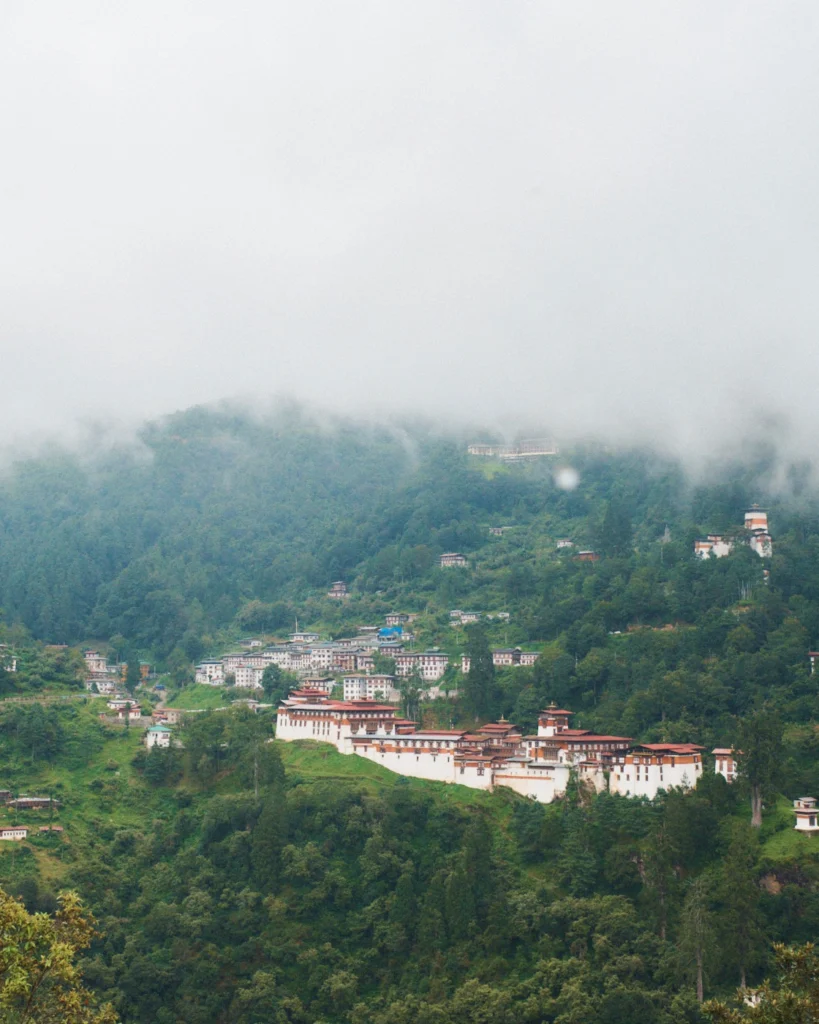 A long dzong on a forested ridge above a small town, with low cloud over the hills