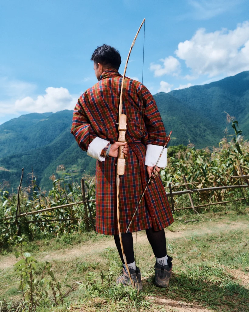 A man in a traditional gho holding a bow on a hillside above terraced fields