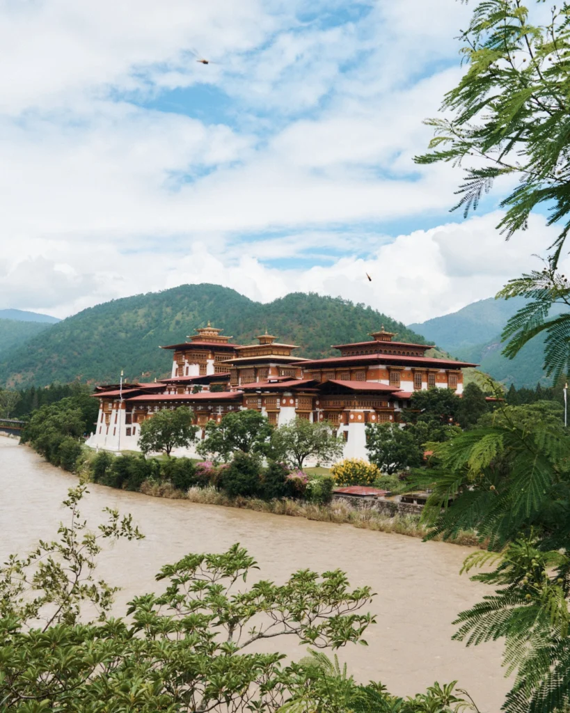 Punakha Dzong beside a wide river, with forested hills and clouds behind