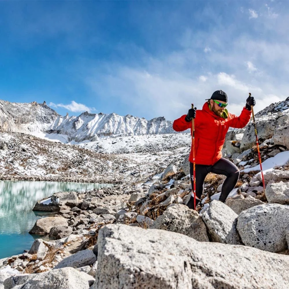 A trekker in a red jacket with walking poles on rocks beside a glacial lake, below snow-covered peaks
