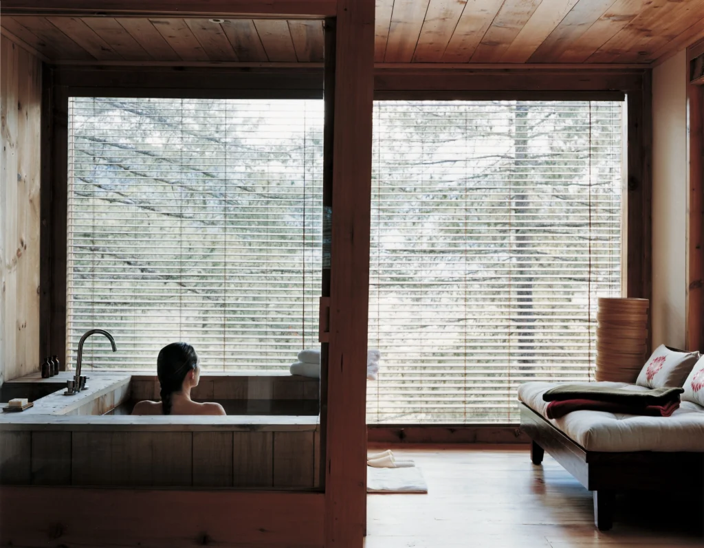 A guest relaxing in a wooden bathhouse with tall windows onto a forest